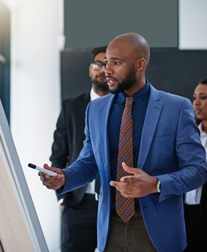 A man in a blue suit presents at a whiteboard with a marker, while two colleagues stand behind him, listening attentively in a modern office setting.