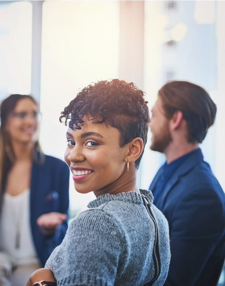 A woman with short curly hair and a nose ring smiles at the camera while two people talk in the blurred background in a bright office setting.