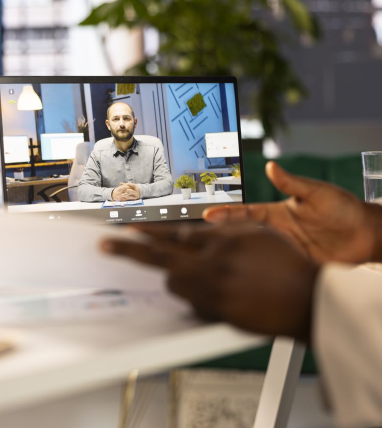A person participates in a video call on a laptop, showing a man sitting at a desk with office decor in the background. The foreground shows hands holding papers near the screen.