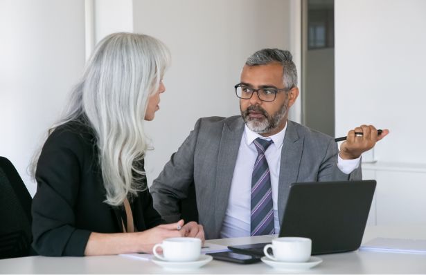 Two businesspeople sit at a desk with a laptop and coffee cups, engaged in conversation. The man gestures with a pen while the woman listens attentively. Both are dressed in professional attire.