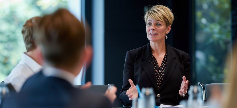 A woman in a black blazer speaks and gestures with her hands during a meeting, while other people listen around a table in a modern office.