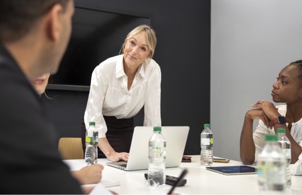 Four people sit around a conference table with water bottles. A woman stands, leaning on the table and looking at a man in the foreground, suggesting a business meeting or discussion.