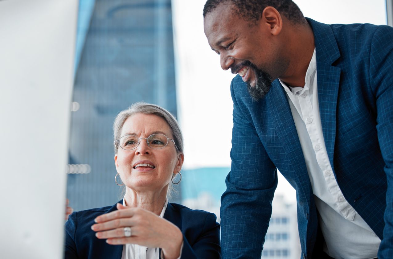 Two business professionals in suits discussing something while looking at a computer screen in a modern office with large windows. The woman gestures as she speaks, and the man beside her is smiling.