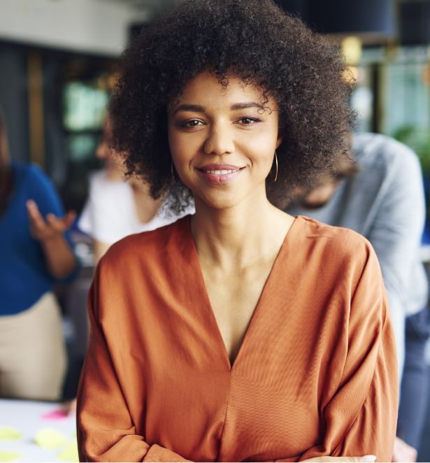 A woman with curly hair, wearing an orange blouse, smiles gently while standing indoors in a bright, modern space. Other people and blurred office elements are visible in the background.