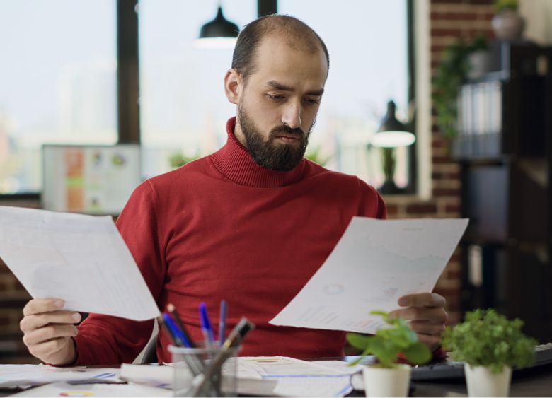 A man with a beard wearing a red sweater sits at a desk, looking at documents in both hands. The desk has papers, pens, and a small plant. There are shelves and a window in the background.