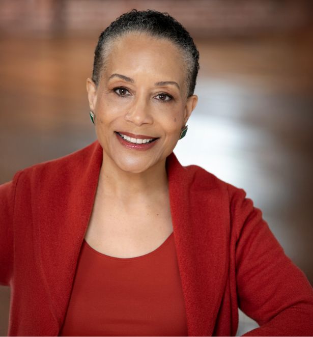 Smiling woman with short hair wears a red top and matching jacket, sitting in a softly lit room with a blurred, brown background.