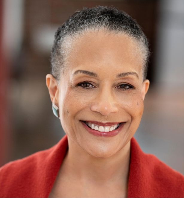A smiling woman with short curly gray hair wears a red coat and turquoise earrings, standing in front of a softly blurred background.